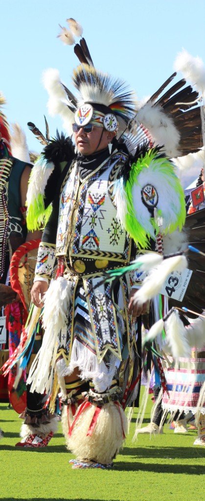 Snow Mountain Paiute Pow Wow Grand Entry as a man in sunglasses adorned in Prairie Hen colorful outfit follows the circle as seen in Powwows: Duck Valley; Elko; Ely; Fallon; Fort Mojave; Goshute; Moapa; Muckleshoot; Nevada Day; Numaga; Red Dress; Sacred Visions; Stewart; Sycuan; UNLV; Univ. of Nev. Reno; and Walker River.